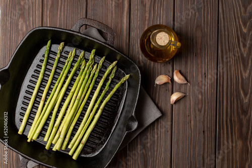 there is a grill pan on a wooden table, next to it is fresh asparagus, garlic cloves and olive oil in a glass bottle, top view, open space