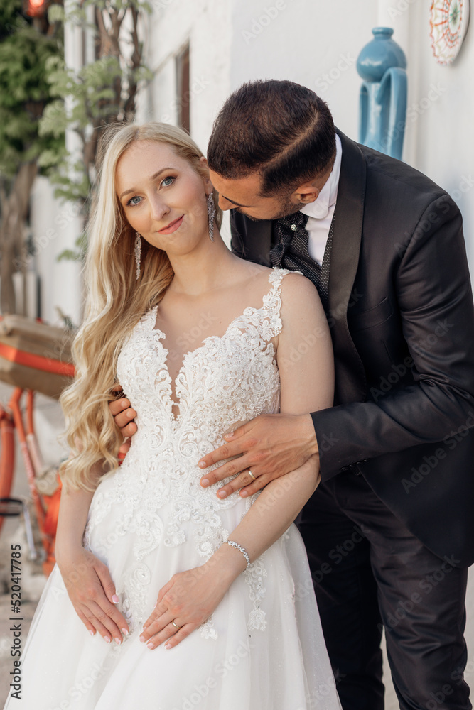 Naklejka premium Portrait of wedding couple standing on street. Young woman looking at camera, groom in suit hugging bride from behind. 