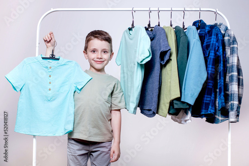 A little stylish preschool boy staiding near the clothes rack, holding T-shirt and demonstrates them