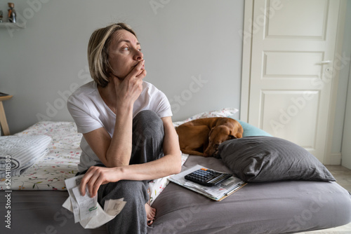 Single depressed mature woman widow loses life sense and motivation. Middle-aged blonde female wearing nightwear sits on bed holding hand on chin looking in distance and waiting for help closeup