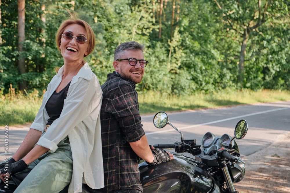 Middle age couple talking and having fun, sitting on a motorcycle, traveling together on a forest road