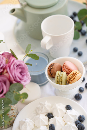 Macarons in white bowl, candles, meringue, teapot and pink roses on the table, tea party with sweets