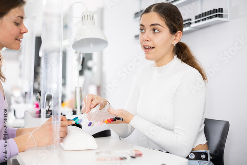 Smiling young female manicurist showing palette of nail varnishes to woman client in beauty salon..