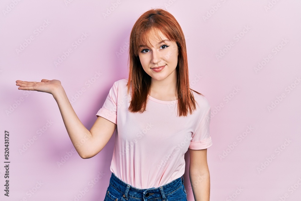 Redhead young woman wearing casual pink t shirt smiling cheerful presenting and pointing with palm of hand looking at the camera. Redhead young woman wearing casual pink t shirt smiling cheerful presenting and pointing with palm of hand looking at the camera.