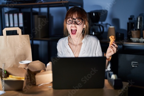 Young beautiful woman working using computer laptop and eating delivery food angry and mad screaming frustrated and furious  shouting with anger. rage and aggressive concept.
