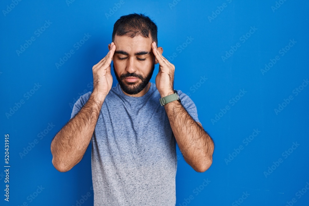 Naklejka premium Middle east man with beard standing over blue background with hand on head, headache because stress. suffering migraine.