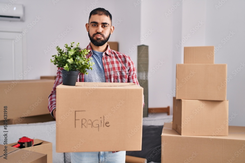 Middle east man with beard moving to a new home holding cardboard box puffing cheeks with funny face. mouth inflated with air, catching air. Middle east man with beard moving to a new home holding cardboard box puffing cheeks with funny face. mouth inflated with air, catching air.