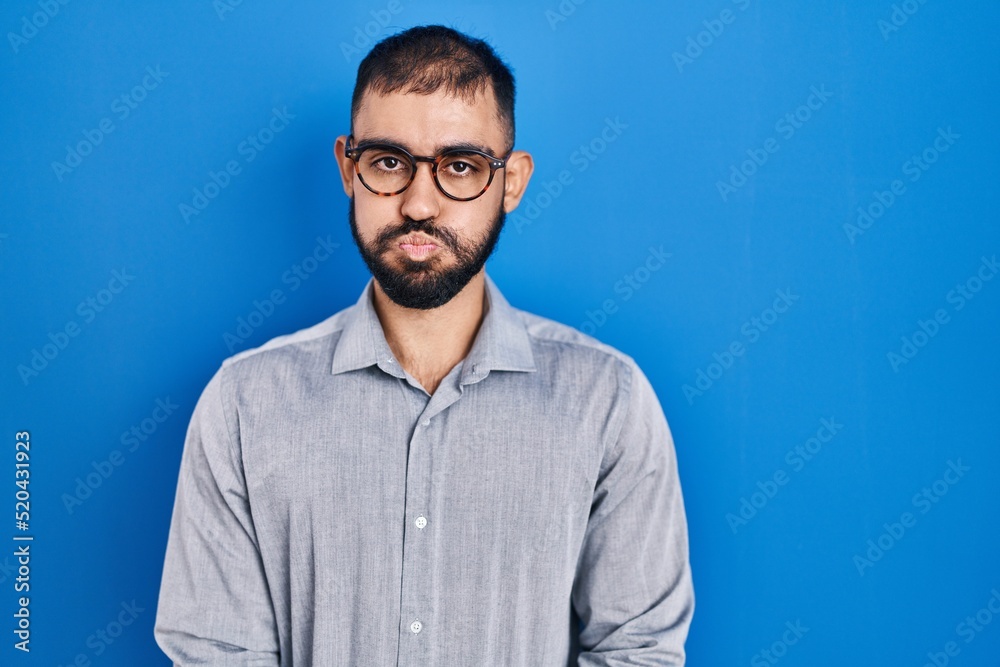 Middle east man with beard standing over blue background puffing cheeks with funny face. mouth inflated with air, crazy expression. Middle east man with beard standing over blue background puffing cheeks with funny face. mouth inflated with air, crazy expression.