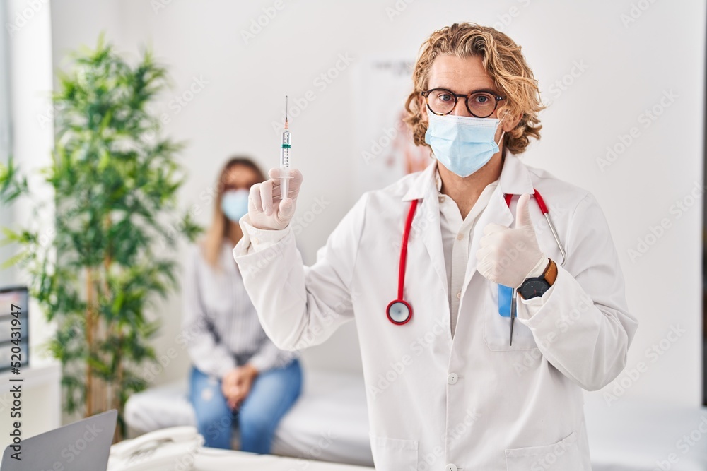Blond man wearing doctor uniform and medical mask holding syringe smiling happy and positive, thumb up doing excellent and approval sign Blond man wearing doctor uniform and medical mask holding syringe smiling happy and positive, thumb up doing excellent and approval sign
