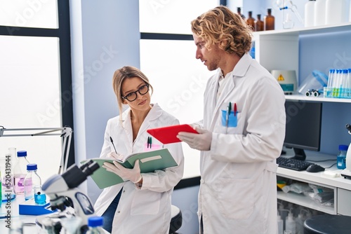 Man and woman wearing scientist uniform writing on notebook using touchpad at laboratory