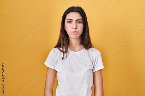 Young beautiful woman standing over yellow background puffing cheeks with funny face. mouth inflated with air, crazy expression.
