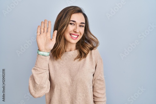 Young woman standing over isolated background waiving saying hello happy and smiling, friendly welcome gesture