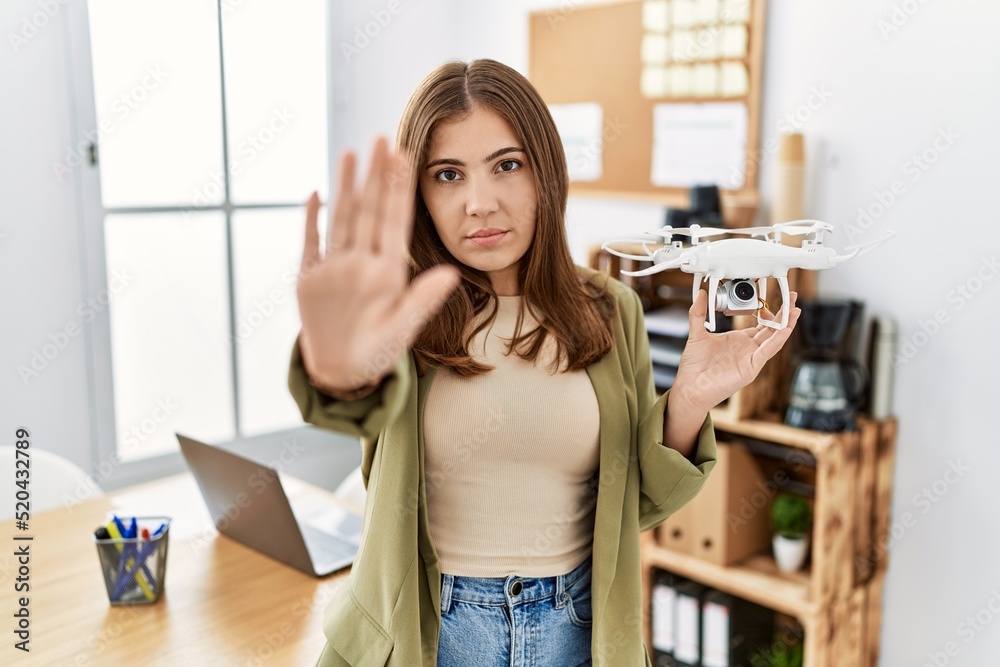 Young brunette woman holding drone at architect office with open hand doing stop sign with serious and confident expression, defense gesture Young brunette woman holding drone at architect office with open hand doing stop sign with serious and confident expression, defense gesture