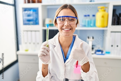 Young blonde woman working at scientist laboratory holding sample smiling happy and positive  thumb up doing excellent and approval sign