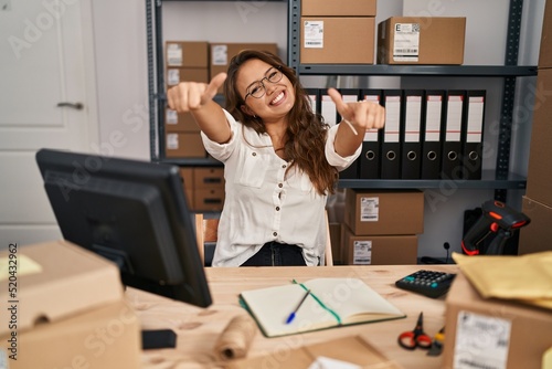 Young hispanic woman working at small business ecommerce approving doing positive gesture with hand, thumbs up smiling and happy for success. winner gesture.