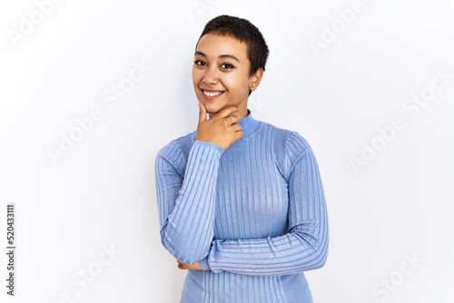 Young hispanic woman with short hair standing over isolated background looking confident at the camera with smile with crossed arms and hand raised on chin. thinking positive.