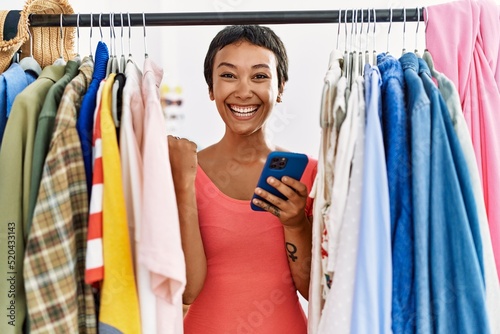 Young hispanic woman with short hair searching clothes on clothing rack using smartphone screaming proud, celebrating victory and success very excited with raised arms