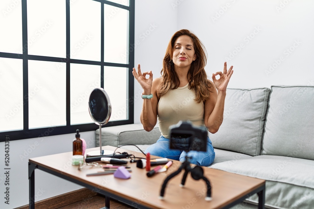 Middle age hispanic woman recording make up tutorial with smartphone at home relax and smiling with eyes closed doing meditation gesture with fingers. yoga concept.