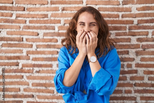 Beautiful brunette woman standing over bricks wall laughing and embarrassed giggle covering mouth with hands, gossip and scandal concept