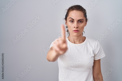 Beautiful brunette woman standing over isolated background pointing with finger up and angry expression, showing no gesture