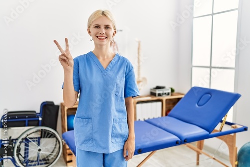 Beautiful caucasian physiotherapist woman working at pain recovery clinic showing and pointing up with fingers number two while smiling confident and happy.