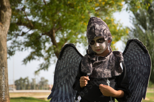 Boy in make up with white face and black eyes, dressed as angel of death with wings and serious look celebrating halloween in an outdoor park. Autumn concept, trick or treat, fear, terror.