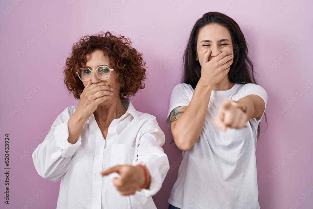 Hispanic mother and daughter wearing casual white t shirt over pink background laughing at you, pointing finger to the camera with hand over mouth, shame expression Hispanic mother and daughter wearing casual white t shirt over pink background laughing at you, pointing finger to the camera with hand over mouth, shame expression