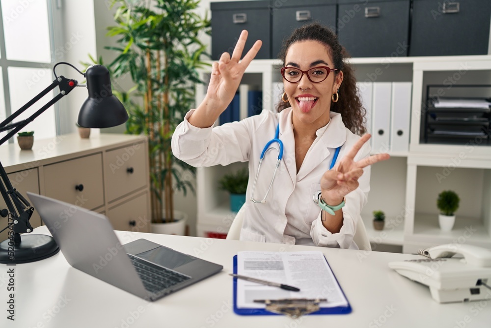 Young hispanic woman wearing doctor uniform and stethoscope smiling with tongue out showing fingers of both hands doing victory sign. number two. Young hispanic woman wearing doctor uniform and stethoscope smiling with tongue out showing fingers of both hands doing victory sign. number two.