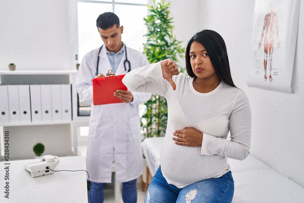 Obraz premium Young hispanic mother expecting a baby at the doctor with angry face, negative sign showing dislike with thumbs down, rejection concept