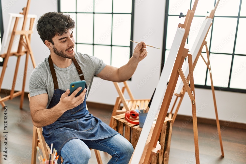 Young hispanic artist man smiling happy painting and using smartphone at art studio. Young hispanic artist man smiling happy painting and using smartphone at art studio.