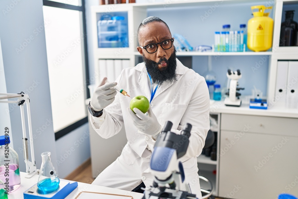 African american man working at scientist laboratory with apple making fish face with mouth and squinting eyes, crazy and comical. African american man working at scientist laboratory with apple making fish face with mouth and squinting eyes, crazy and comical.