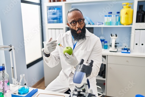 African american man working at scientist laboratory with apple making fish face with mouth and squinting eyes  crazy and comical.