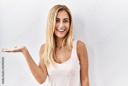 Blonde beautiful young woman standing over white isolated background smiling cheerful presenting and pointing with palm of hand looking at the camera.