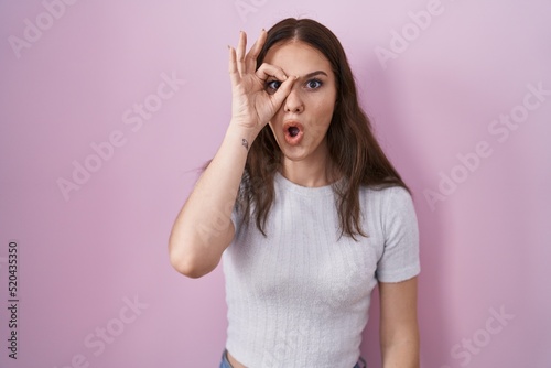Young hispanic girl standing over pink background doing ok gesture shocked with surprised face, eye looking through fingers. unbelieving expression.