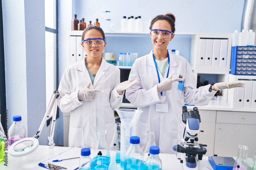 Young mother and daughter at scientist laboratory amazed and smiling to the camera while presenting with hand and pointing with finger. Young mother and daughter at scientist laboratory amazed and smiling to the camera while presenting with hand and pointing with finger.