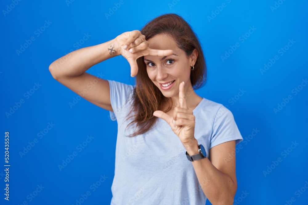 Brunette woman standing over blue background smiling making frame with hands and fingers with happy face. creativity and photography concept. Brunette woman standing over blue background smiling making frame with hands and fingers with happy face. creativity and photography concept.