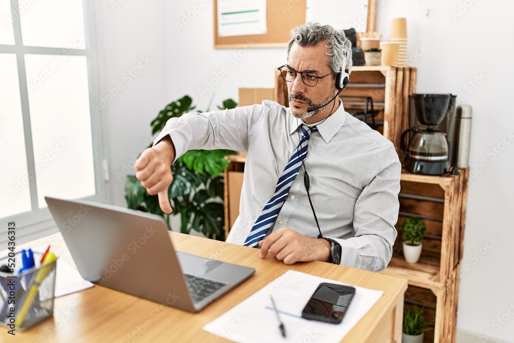 Fototapeta premium Middle age hispanic business man working at the office wearing operator headset looking unhappy and angry showing rejection and negative with thumbs down gesture. bad expression.