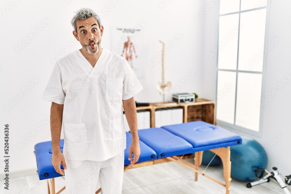 Middle age hispanic therapist man working at pain recovery clinic making fish face with lips, crazy and comical gesture. funny expression. Middle age hispanic therapist man working at pain recovery clinic making fish face with lips, crazy and comical gesture. funny expression.