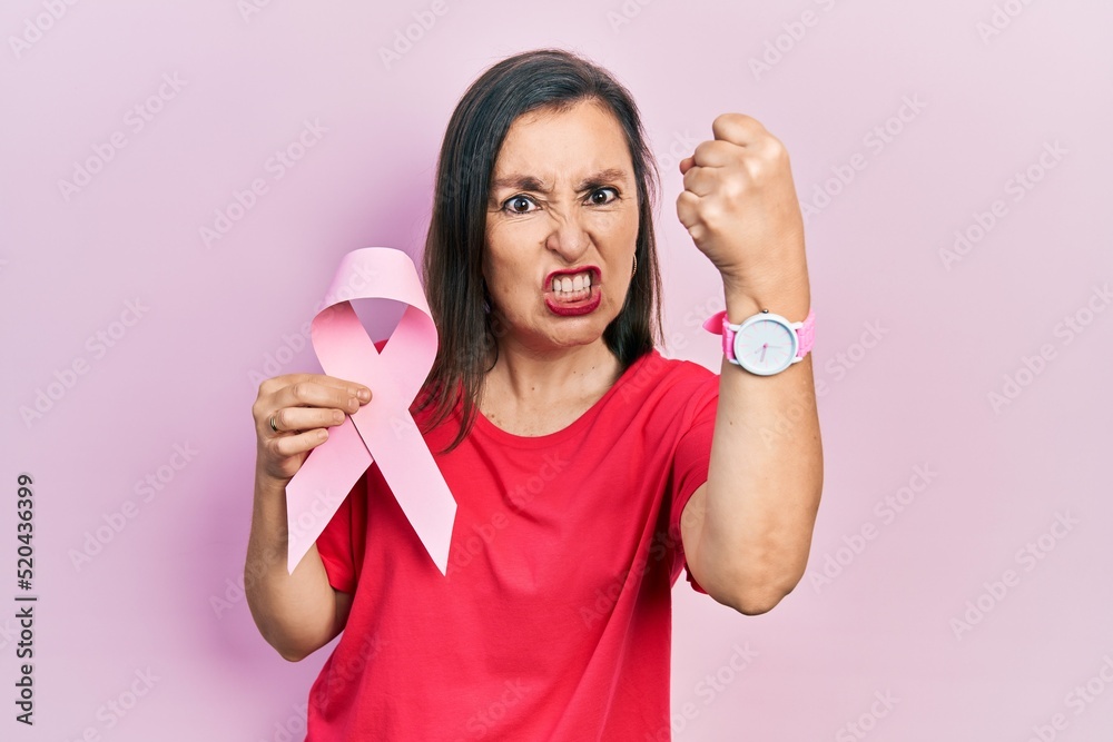 Middle age hispanic woman holding pink cancer ribbon annoyed and frustrated shouting with anger, yelling crazy with anger and hand raised Middle age hispanic woman holding pink cancer ribbon annoyed and frustrated shouting with anger, yelling crazy with anger and hand raised