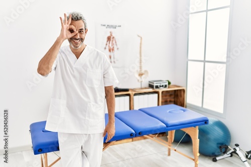 Middle age hispanic therapist man working at pain recovery clinic doing ok gesture with hand smiling  eye looking through fingers with happy face.