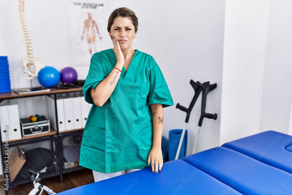 Young hispanic woman wearing physiotherapist uniform standing at clinic touching mouth with hand with painful expression because of toothache or dental illness on teeth. dentist Young hispanic woman wearing physiotherapist uniform standing at clinic touching mouth with hand with painful expression because of toothache or dental illness on teeth. dentist