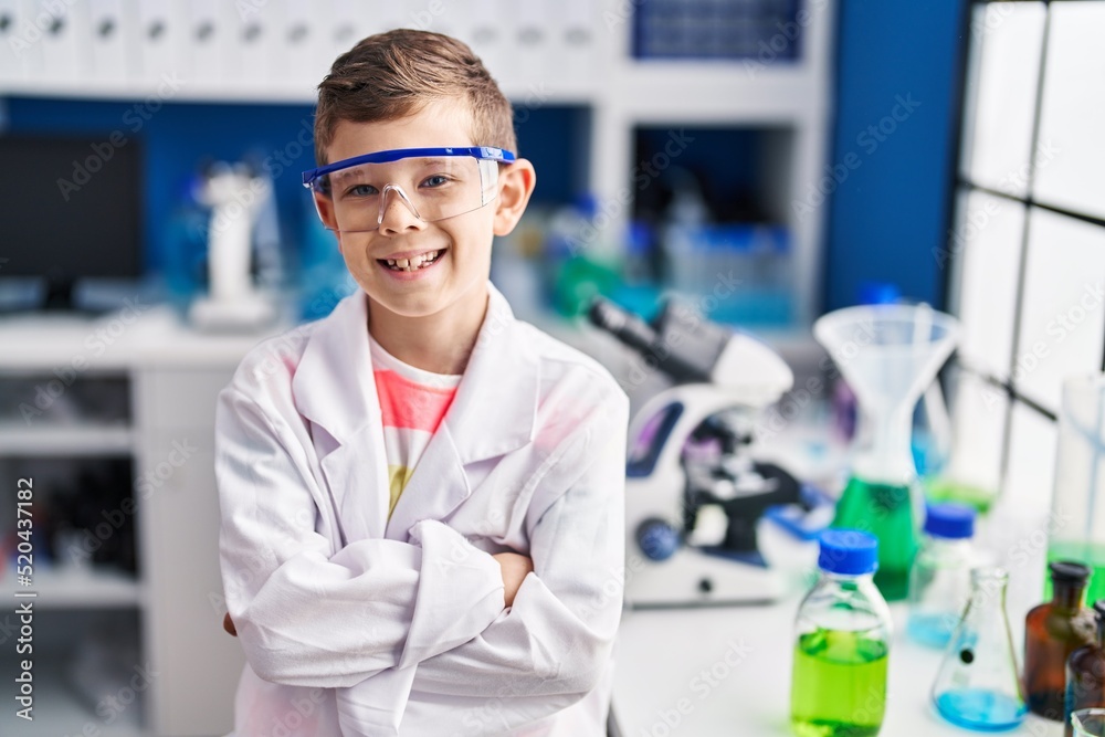 Blond child wearing scientist uniform sitting with arms crossed gesture at laboratory