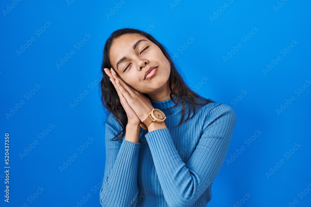Young brazilian woman standing over blue isolated background sleeping tired dreaming and posing with hands together while smiling with closed eyes. Young brazilian woman standing over blue isolated background sleeping tired dreaming and posing with hands together while smiling with closed eyes.