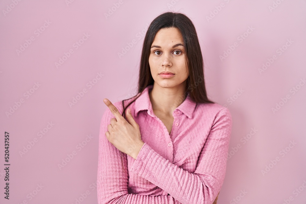 Young hispanic woman standing over pink background pointing with hand finger to the side showing advertisement, serious and calm face Young hispanic woman standing over pink background pointing with hand finger to the side showing advertisement, serious and calm face