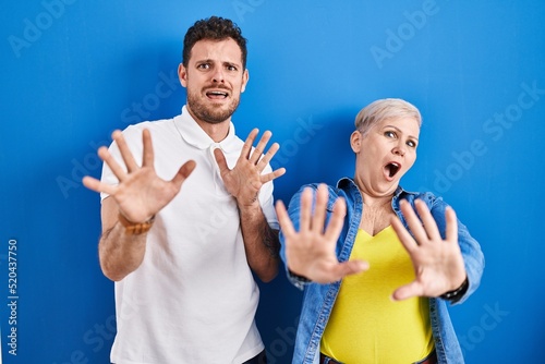 Young brazilian mother and son standing over blue background afraid and terrified with fear expression stop gesture with hands, shouting in shock. panic concept.