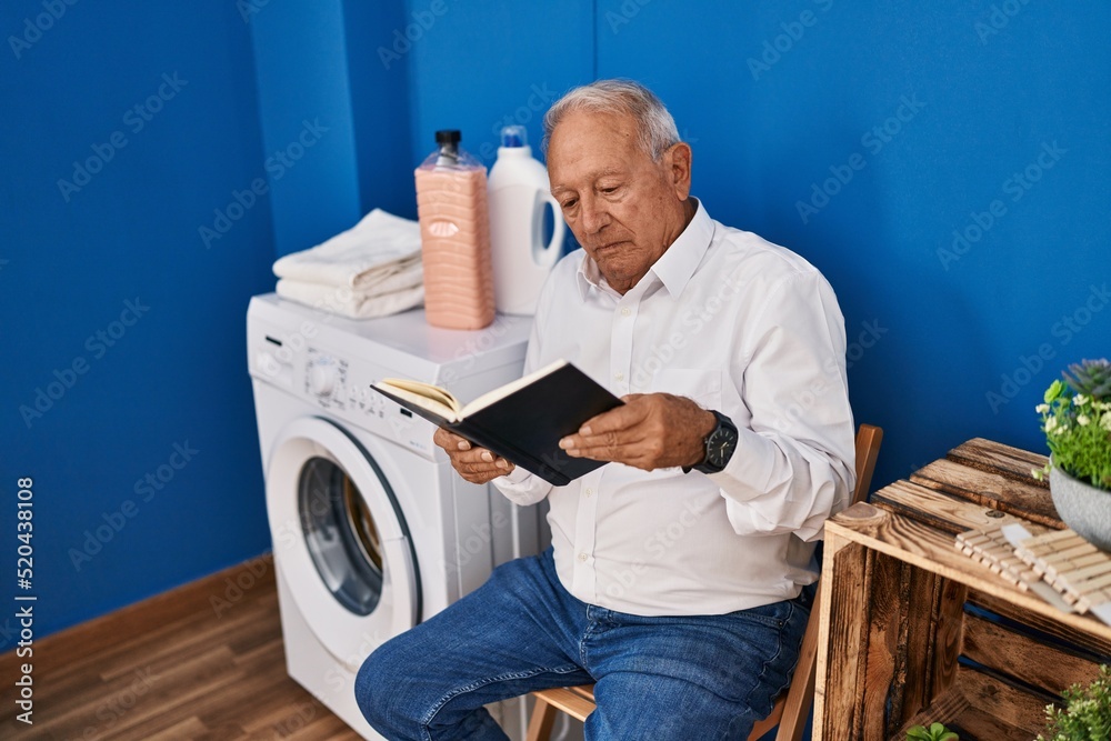 Obraz premium Senior man reading book waiting for washing machine at laundry room