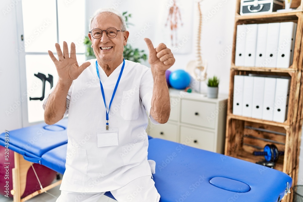 Fototapeta premium Senior physiotherapy man working at pain recovery clinic showing and pointing up with fingers number six while smiling confident and happy.