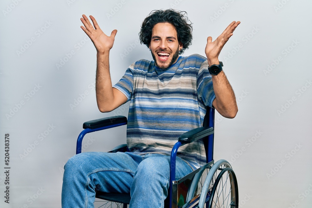 Handsome hispanic man sitting on wheelchair celebrating crazy and amazed for success with arms raised and open eyes screaming excited. winner concept Handsome hispanic man sitting on wheelchair celebrating crazy and amazed for success with arms raised and open eyes screaming excited. winner concept