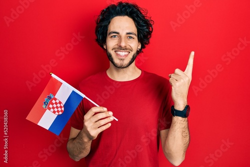 Handsome hispanic man holding croatia flag smiling with an idea or question pointing finger with happy face, number one