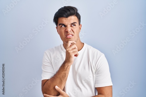 Hispanic man standing over blue background thinking worried about a question, concerned and nervous with hand on chin